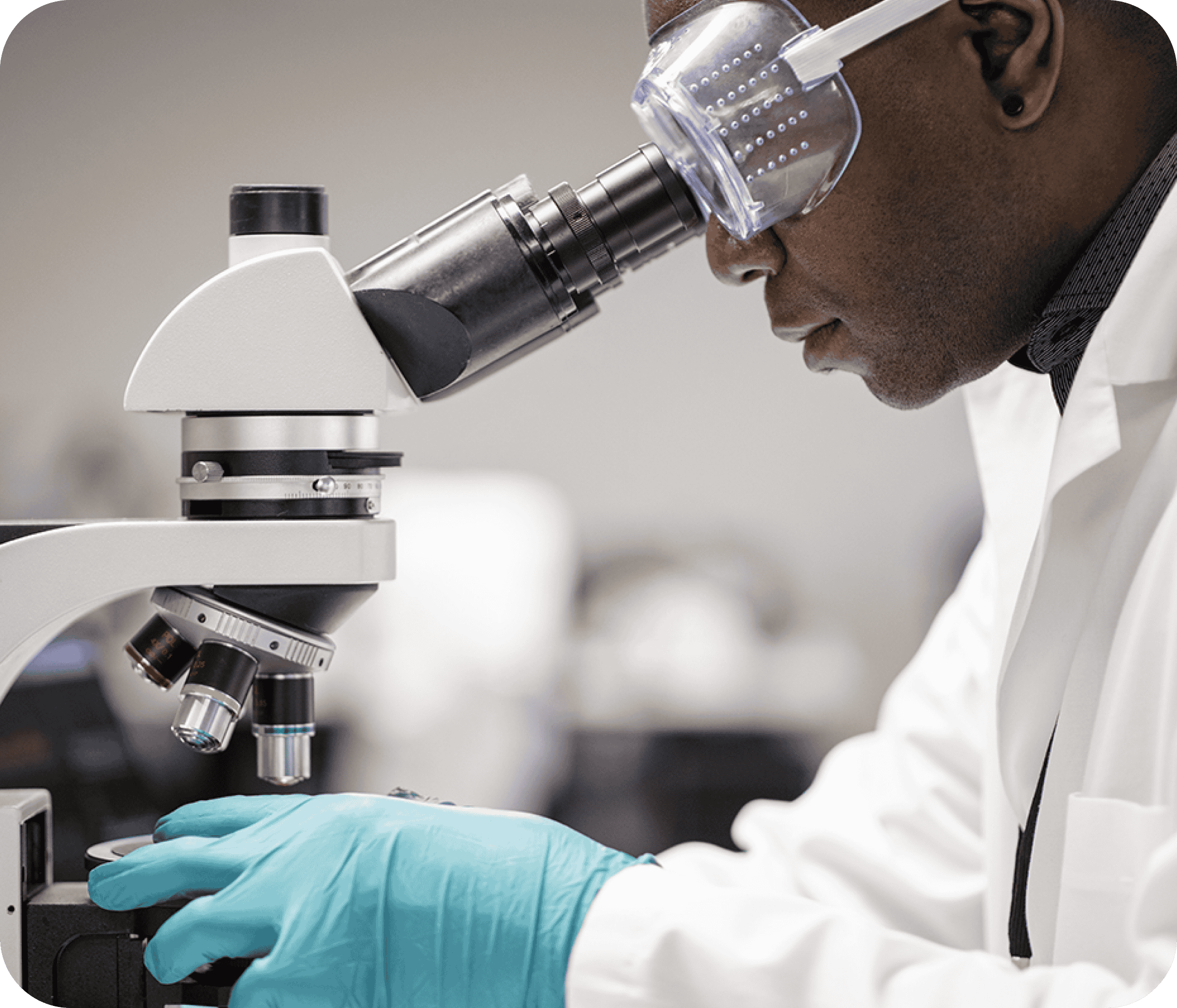 A researcher in a lab coat and wearing gloves intently working in a lab, viewing samples through a microscope.